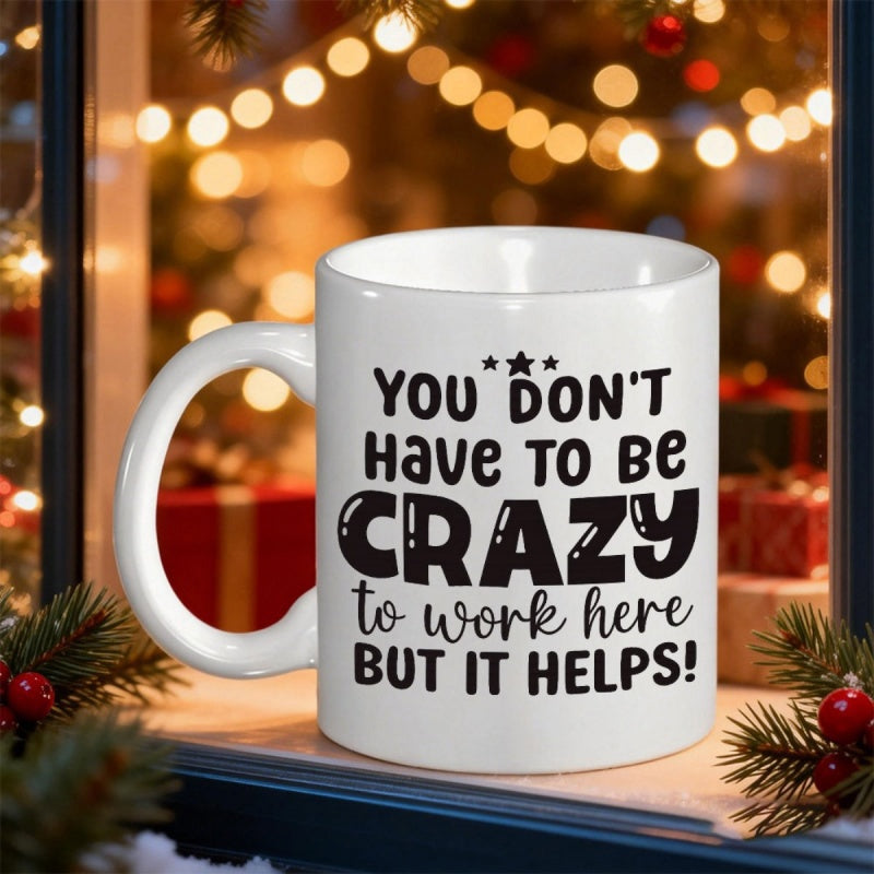 Tailorly White Ceramic 11 oz Mug - You Don't Have To Be Crazy To Work Here But It Helps White Ceramic Cup With Black Text Against Warm Festive Christmas Lights Mug