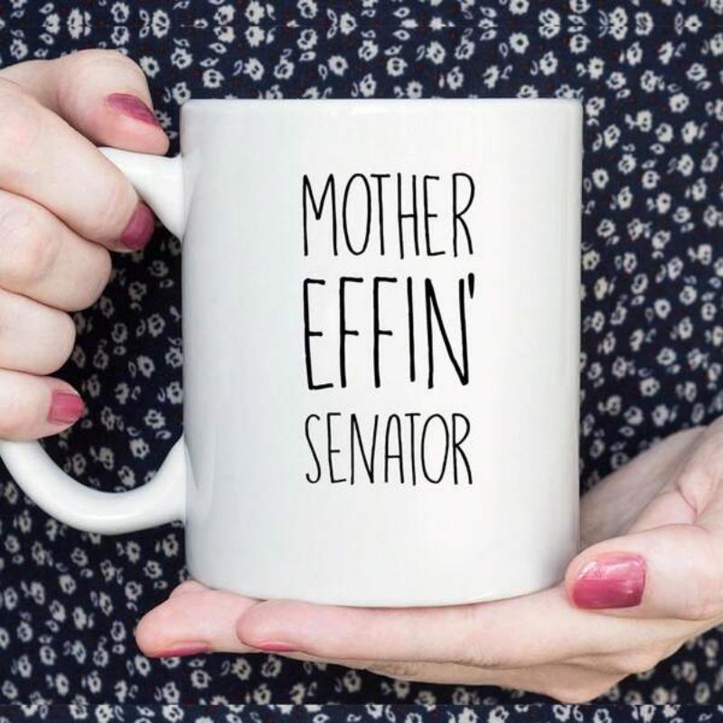 Tailorly White Ceramic 11 oz Mug - Mother Effin Senator Minimalist White Ceramic Vessel Displayed On A Dark Patterned Shirt Background Mug